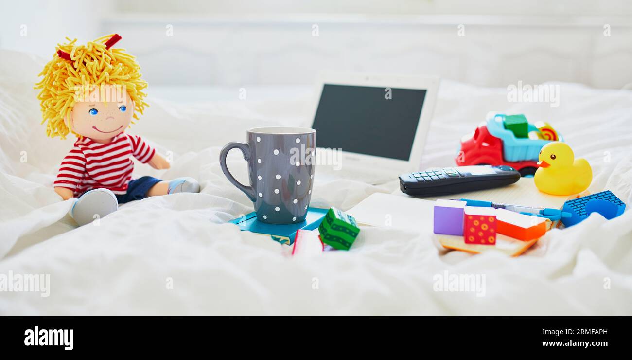 Laptop, cup of coffee, notebook, phone and different toys in bed on clean white linens. Freelance, distance learning or work from home with kids conce Stock Photo