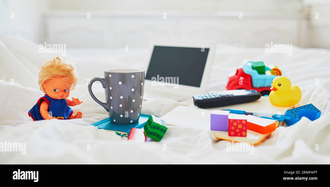 Laptop, cup of coffee, notebook, phone and different toys in bed on clean white linens. Freelance, distance learning or work from home with kids conce Stock Photo