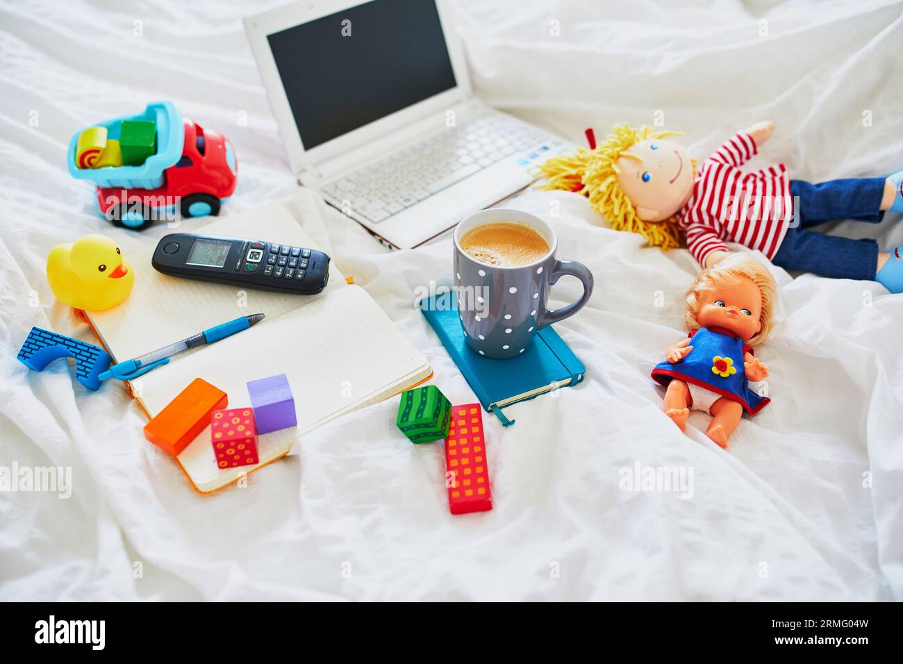 Laptop, cup of coffee, notebook, phone and different toys in bed on clean white linens. Freelance, distance learning or work from home with kids conce Stock Photo