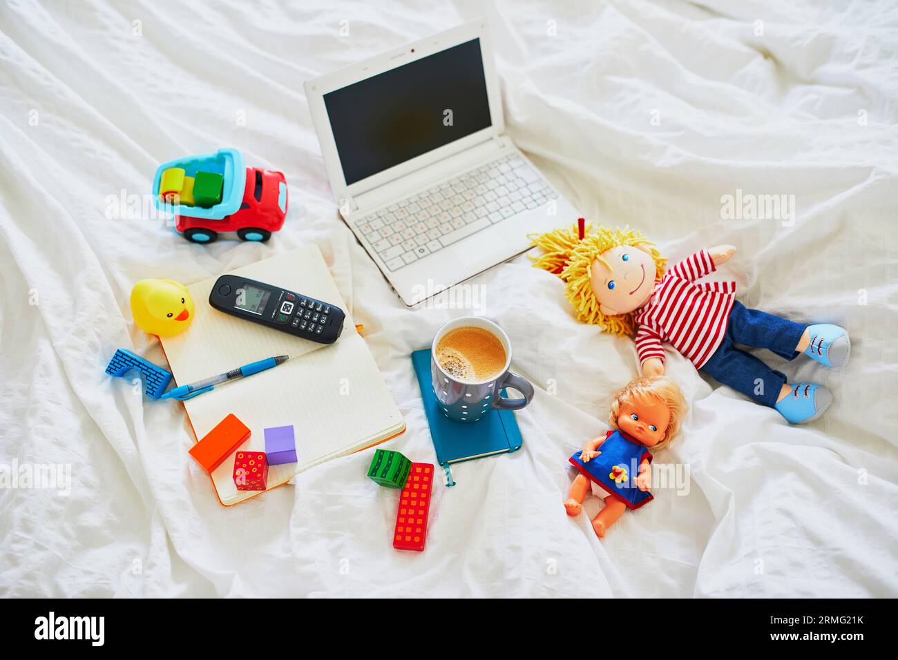 Laptop, cup of coffee, notebook, phone and different toys in bed on clean white linens. Freelance, distance learning or work from home with kids conce Stock Photo