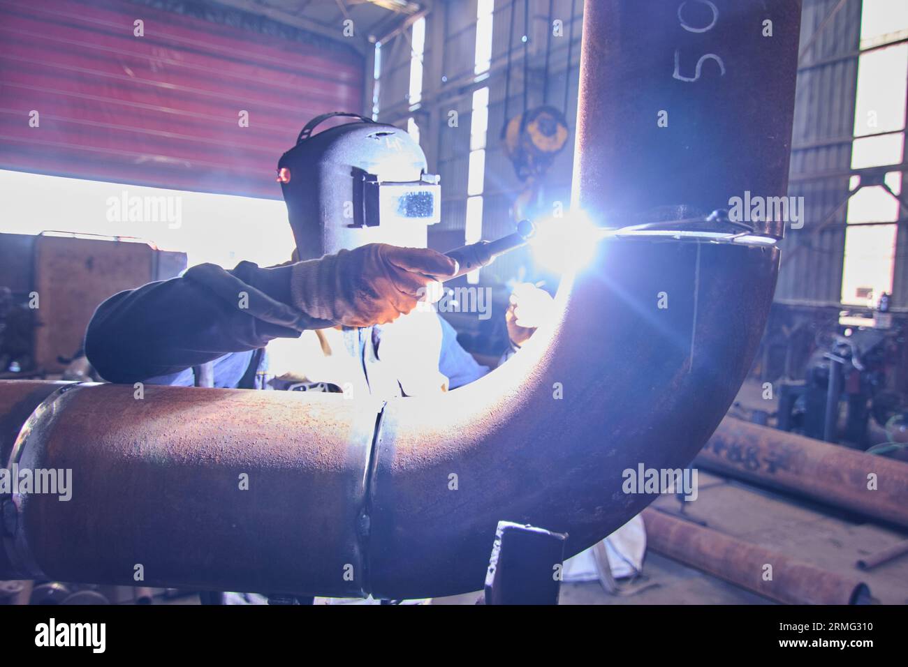 a man in full safety gear like a helmet, visor, and vest. doing welding on a rusted metal pipe. a man using a handheld electrical saw on a metal pipe. Stock Photo