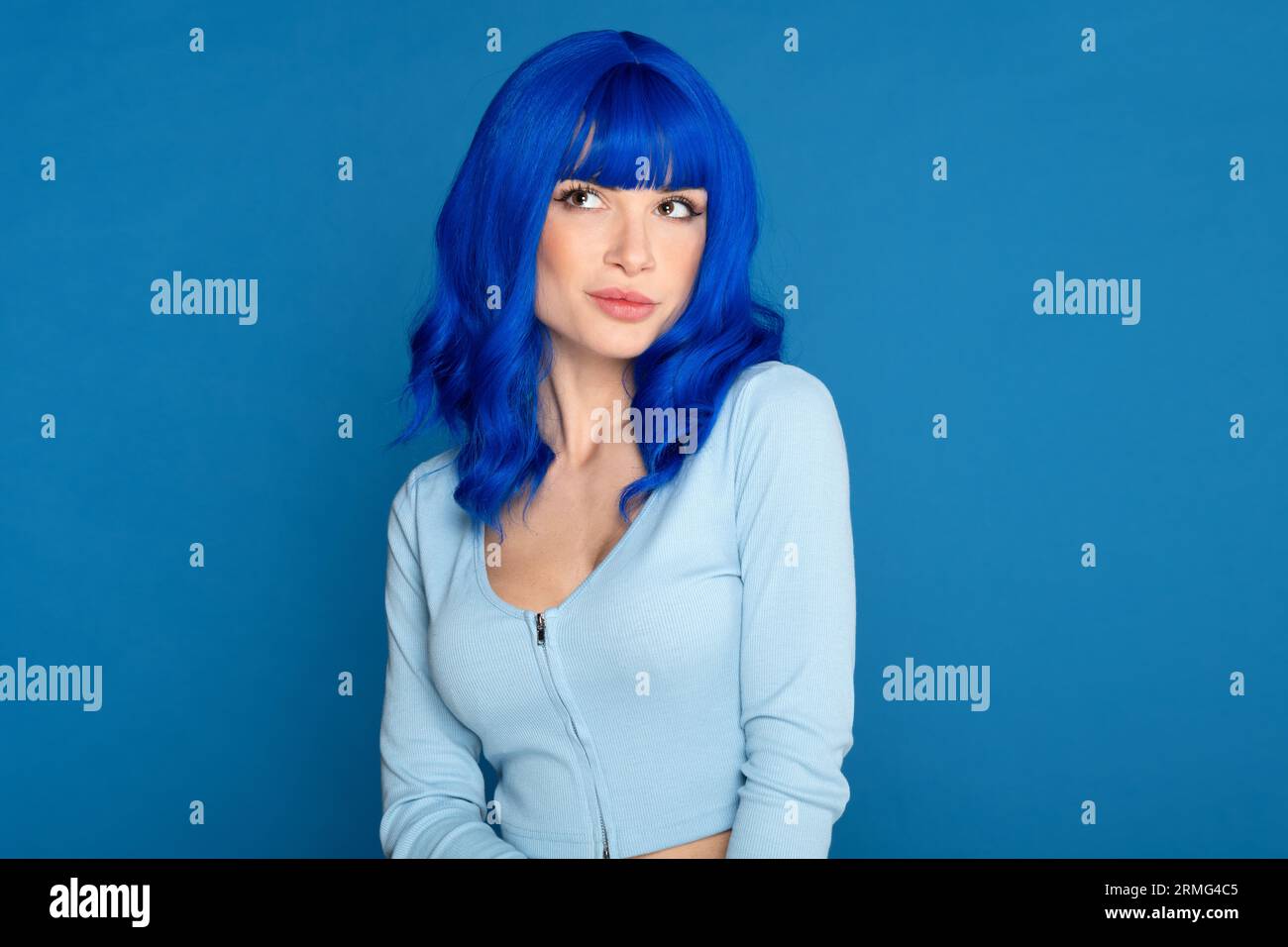 Confident young female model in blue bright wig and casual wear looking away dreamily against blue background in studio Stock Photo