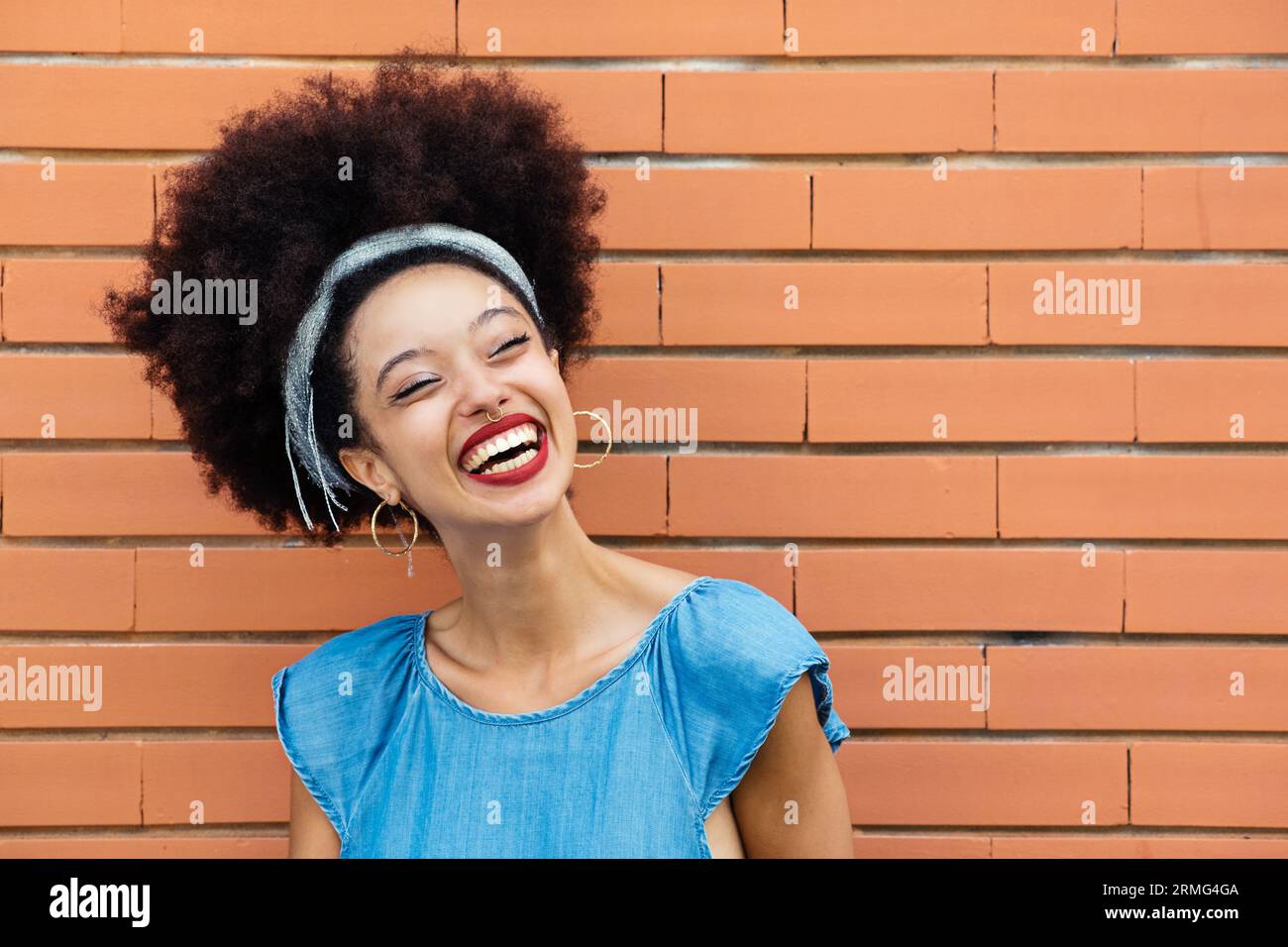 Positive young African American female with Afro hair headband red lips smiling and looking away while standing alone against brown brick wall buildin Stock Photo
