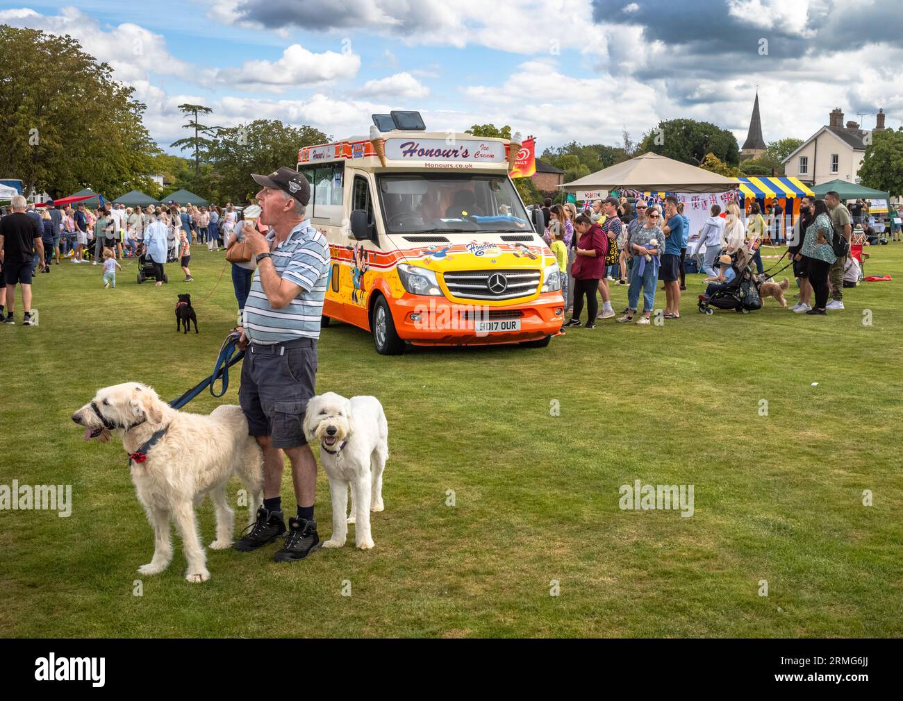 Man with holding two white dogs on leads eats an ice cream next to an ice cream van at  Wisborough Green Village Fete, West Sussex, UK Stock Photo