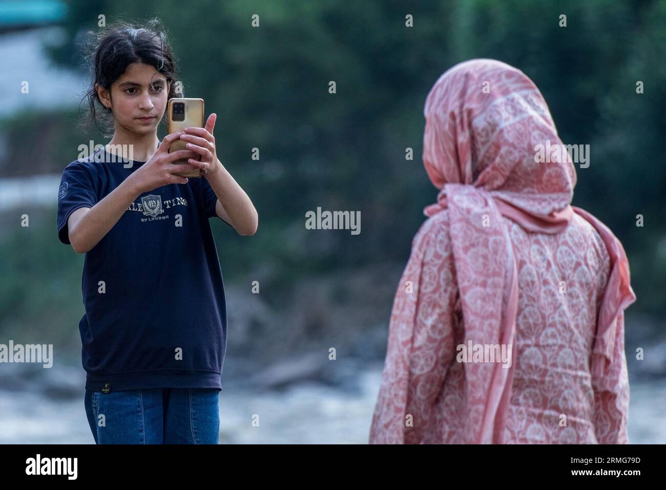 Keran Kupwara, India. 24th Aug, 2023. A girl takes a photo of her mother on the banks of Neelam river or Kishan Ganga that has divided Kashmir into two parts controlled by nuclear rivals India and Pakistan. The river acts as a disputed line of control and flows through a village called Keran from both sides which is located on the northern side of Indian Kashmir's Border district Kupwara some 150kms from Srinagar and 93kms from Muzaffarabad, in the Pakistan side of Kashmir. (Photo by Faisal Bashir/SOPA Images/Sipa USA) Credit: Sipa USA/Alamy Live News Stock Photo