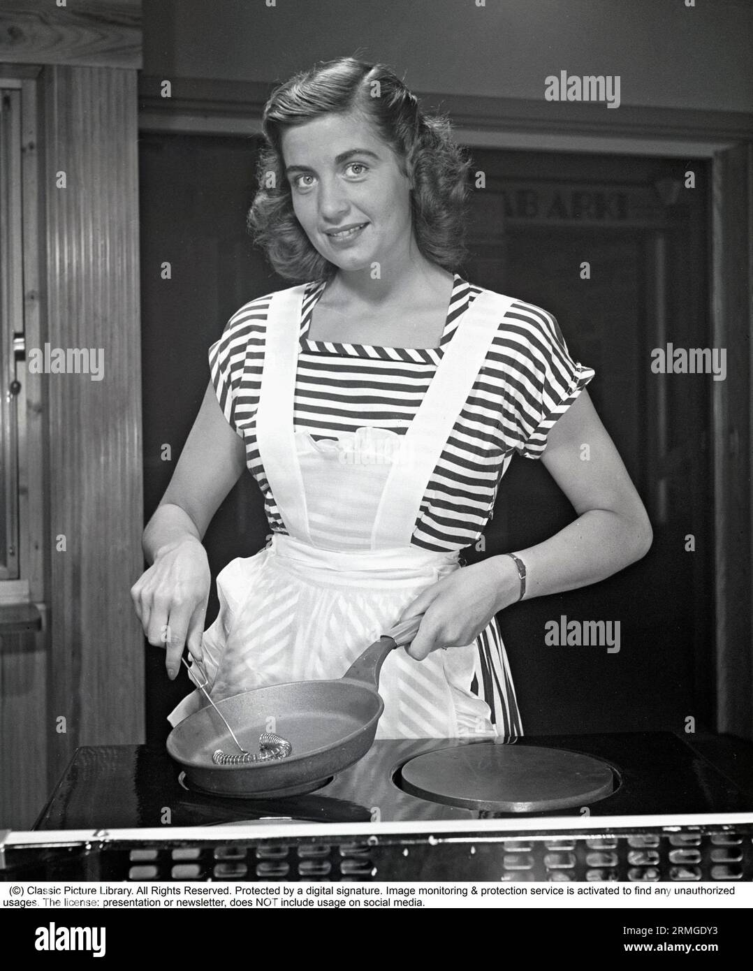 In the kitchen 1940s. A young woman smiles at the camera holding a frying pan and a whisk in her hands. She is wearing a whit apron. Sweden 1948. Kristoffersson ref AL27-8 Stock Photo