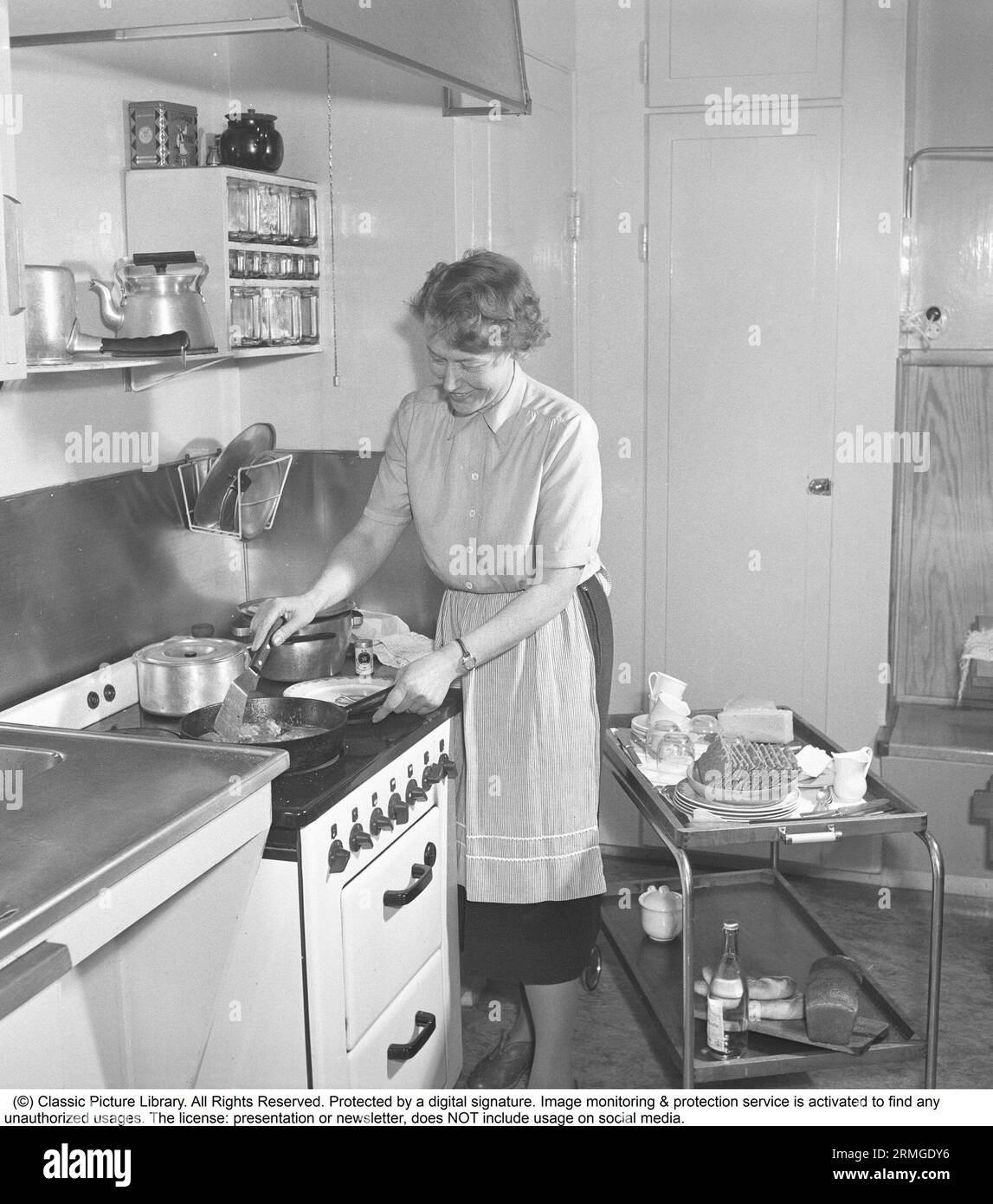 In the kitchen 1950s. Interior of a kitchen and a woman standing at the kitchen cooker frying something in the frying pan. She is wearing a apron.  Sweden 1952. Kristoffersson ref BF82-9 Stock Photo