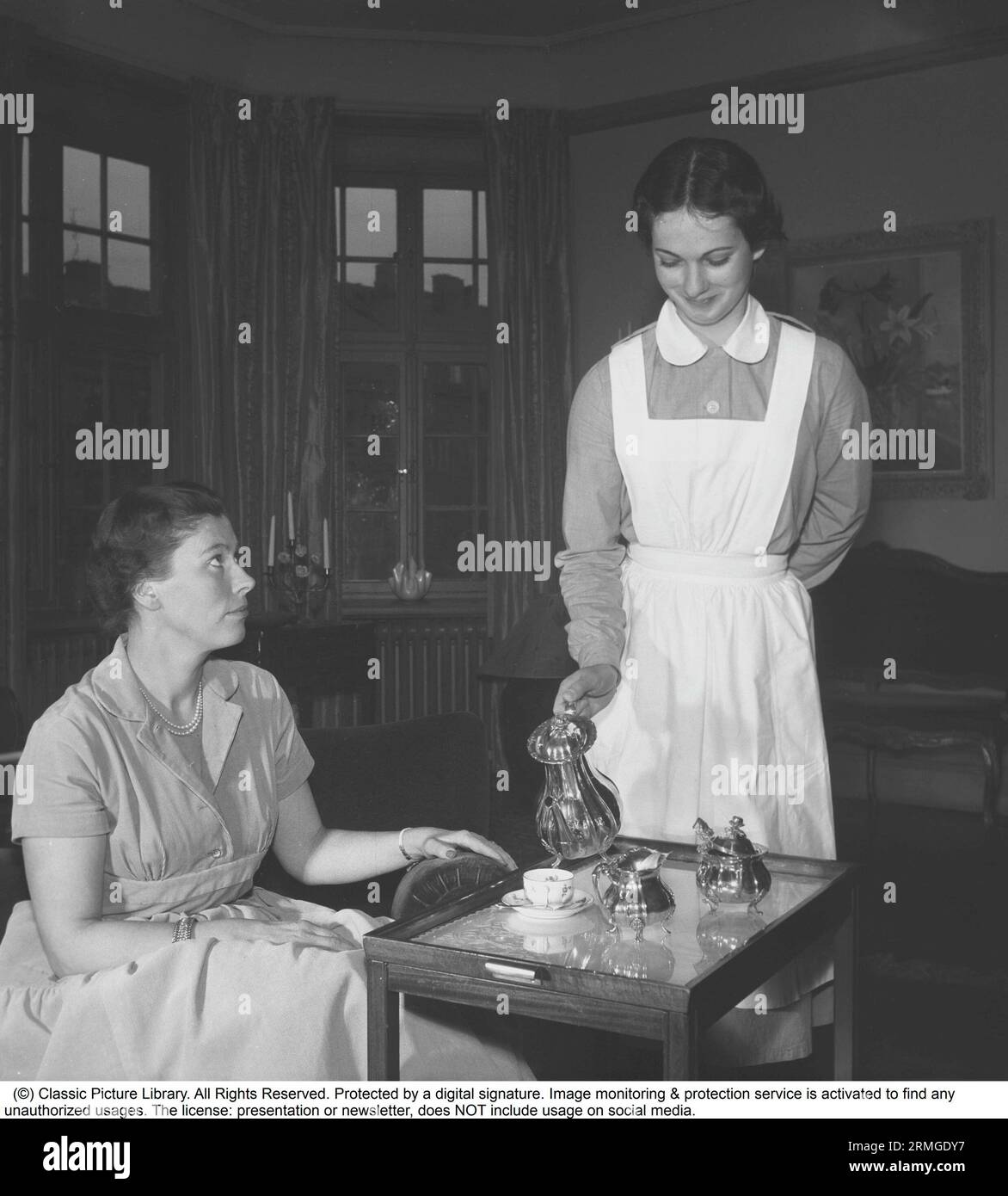 Living in the 1950s. A young woman working as a maid is serving tea or coffee to a woman sitting in the couch. She is neatly dressed in a white apron.    Sweden 1955. Kristoffersson ref BU6-8 Stock Photo