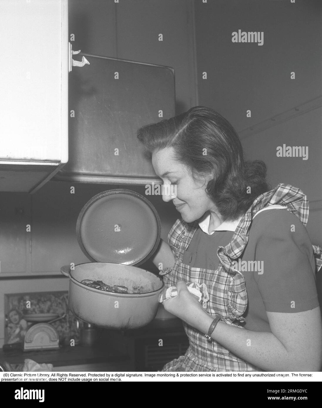 In the kitchen 1940s. Interior of a kitchen and a young woman lifting the lid of a saucepan steaming hot and looks happy with how it turned out. Sweden 1946 Kristoffersson ref CC122-1 Stock Photo