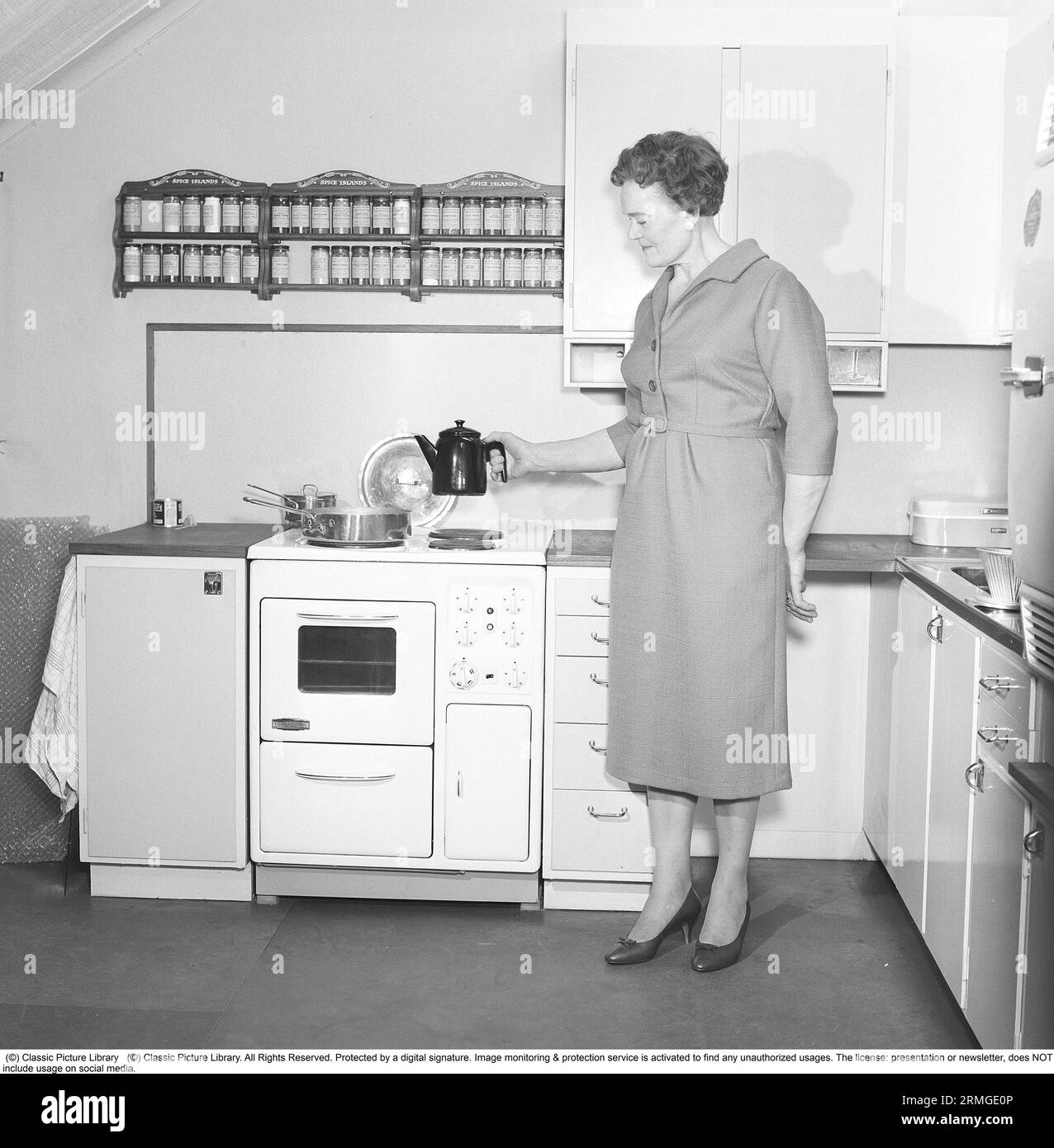 In the 1950s. A woman in a typical 1950s kitchen with wooden cabinets and a set of spices on shelfes above the cooker. She is holding a coffee pot in the typical 50s enamelled look. Sweden 1959. Kristoffersson ref CL8-9 Stock Photo