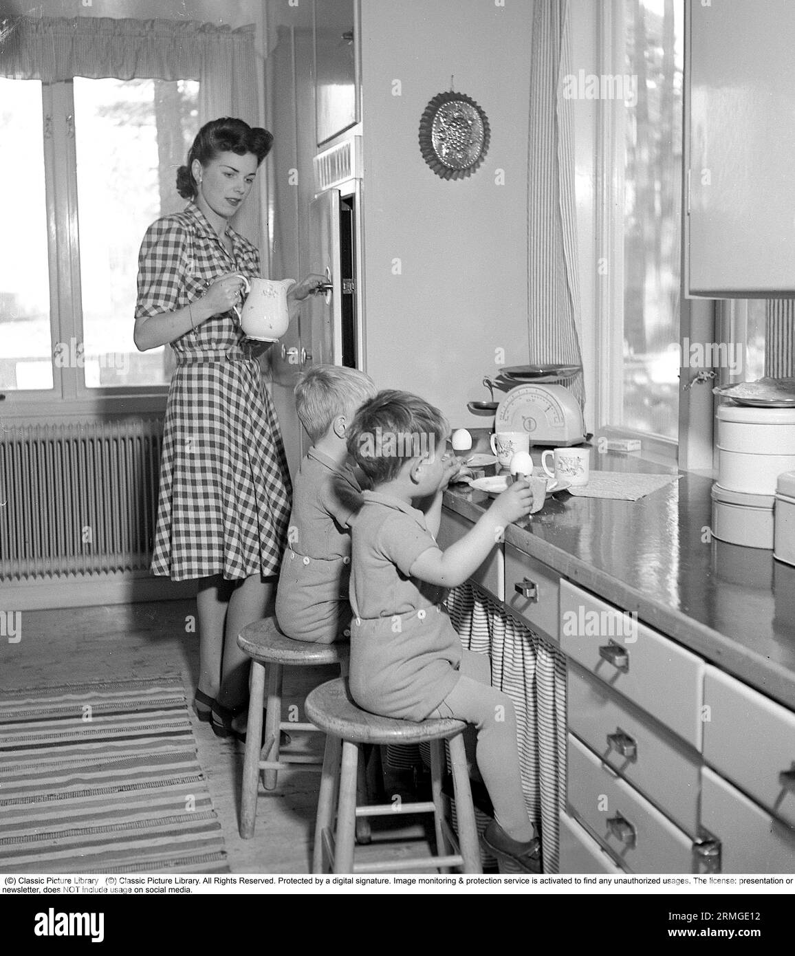 A young mother with her two boys is in the kitchen, and she has served the children eggs, sandwiches and milk, which they eat sitting on stools at the kitchen counter. On the bench is a household scale and tin cans for storing cakes and bread. Sweden 1945. Kristoffersson ref R12-2 Stock Photo