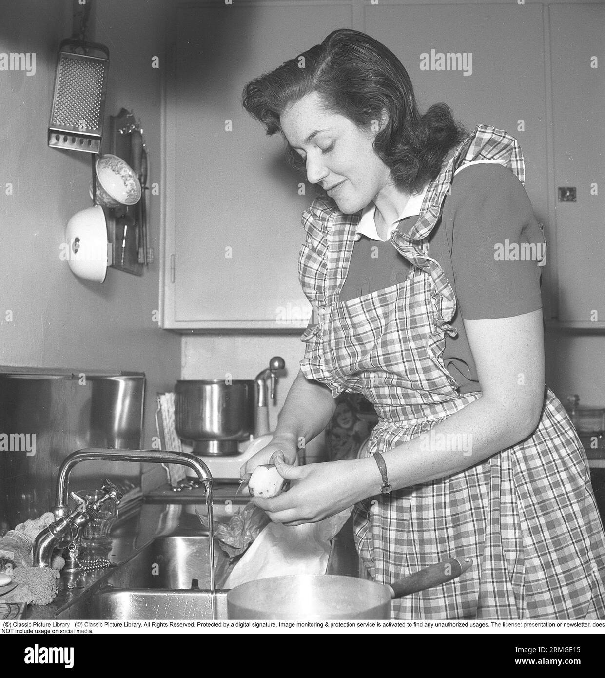 In the 1940s. A woman in a typical 1940s kitchen with the details of the interior and the objects being very representative of the decade. A household assistant, kitchen machine, is an electrical appliance whose basic function is to mix and process dough and it's seen standing on the bench.    Actually, household assistant is a brand word taken from the Electrolux model called Assistant. Electrolux has registered the word Assistant as a trademark. Unlike, for example, food processors and electric whisks, the processing in an assistant takes place at a significantly lower speed, which is a prer Stock Photo