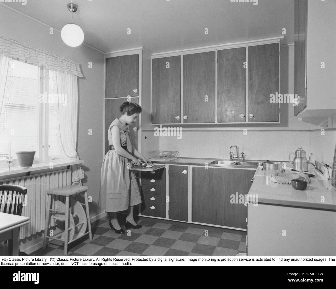 In the 1950s. A woman in a typical 1950s kitchen with wooden cabinets and typical of the decade. She uses the extendable cutting board to slice the sweet bread to eat.  Sweden 1958. Stock Photo