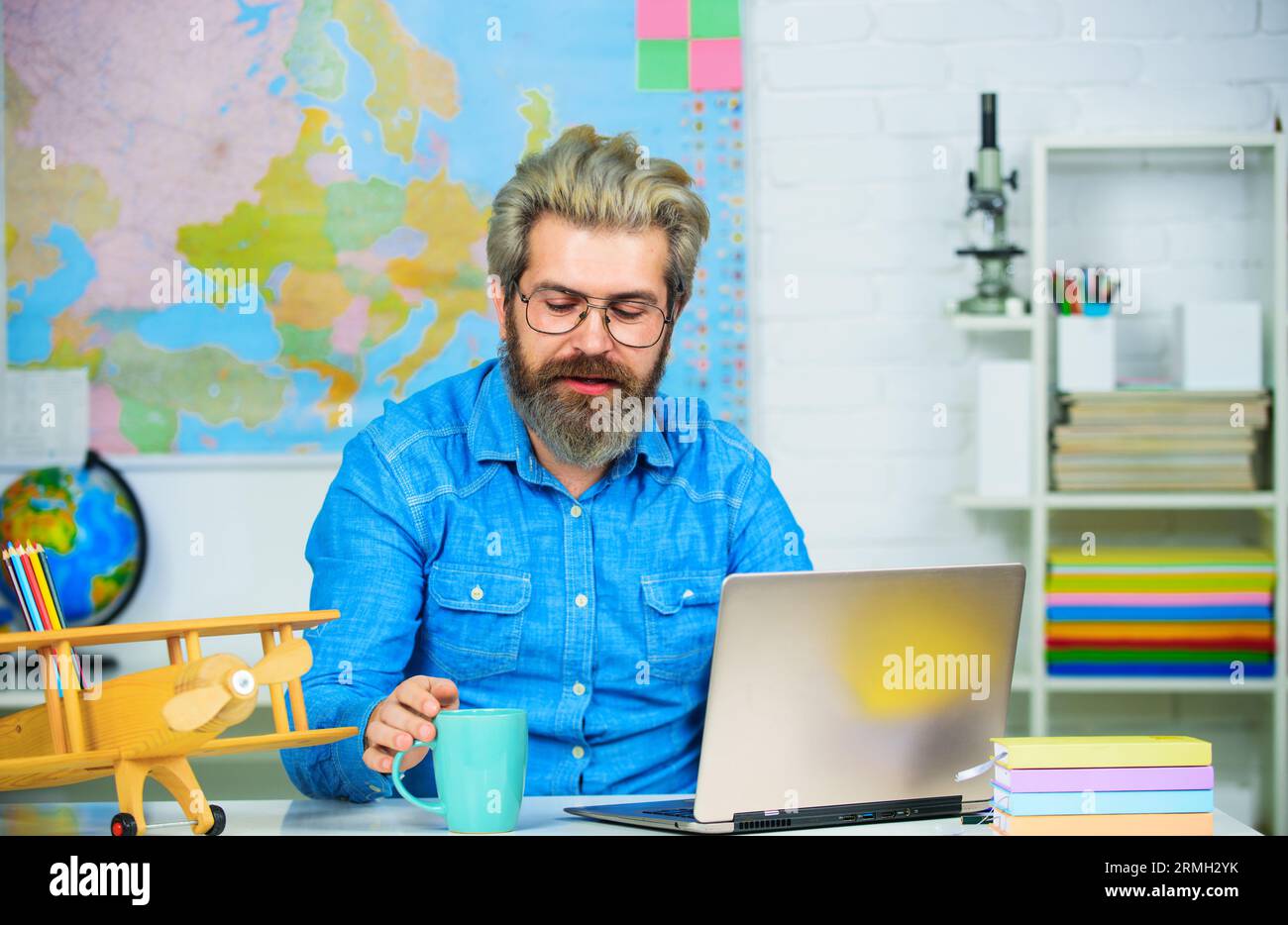 Smiling teacher drinking coffee in classroom. Bearded man drinks coffee