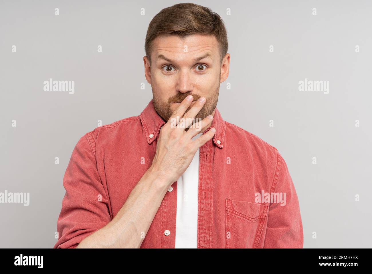 Amazed confused man with raised eyebrows covering mouth with hand looking at camera on studio wall Stock Photo