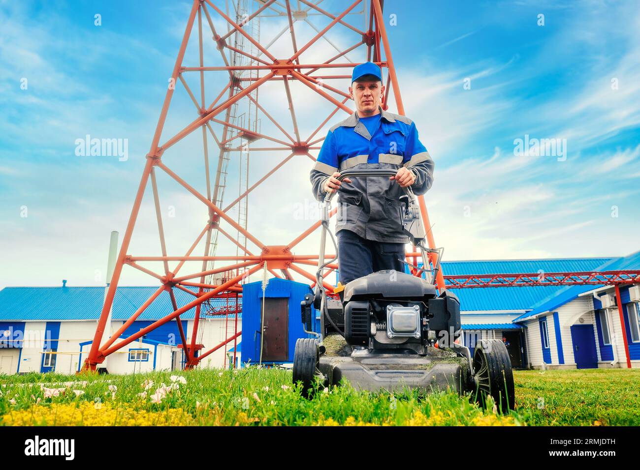 Worker in overalls mows territory of industrial facility with lawn mower. Handyman walks with lawn mower against blue sky. Stock Photo