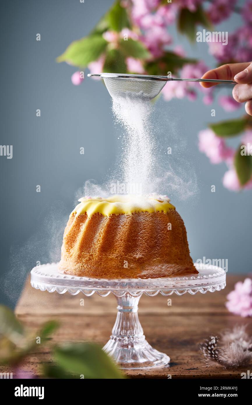 Cropped hand dusting sugar powder on top of the bundt cake. Behind is