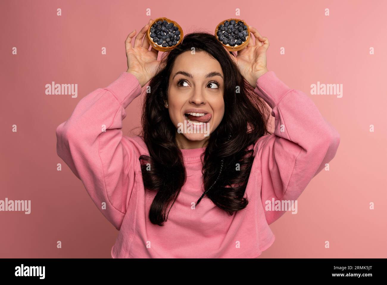 Woman posing for the camera on the pink background with mouth-watering fruit tartlets on her head. Fun and sweet food concept Stock Photo