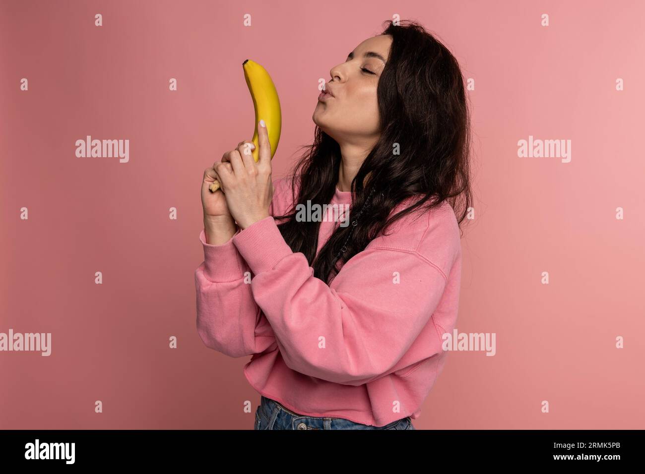 Young dark-haired brunette with a banana gun posing on the pink background during the studio photo shoot. Healthy food concept Stock Photo