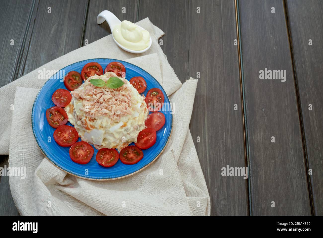 Ensaladilla rusa or russian salad, typical spanish tapa with tuna and mayonnaise on a blue plate decorated with tomato and green leaves on a raffia Stock Photo