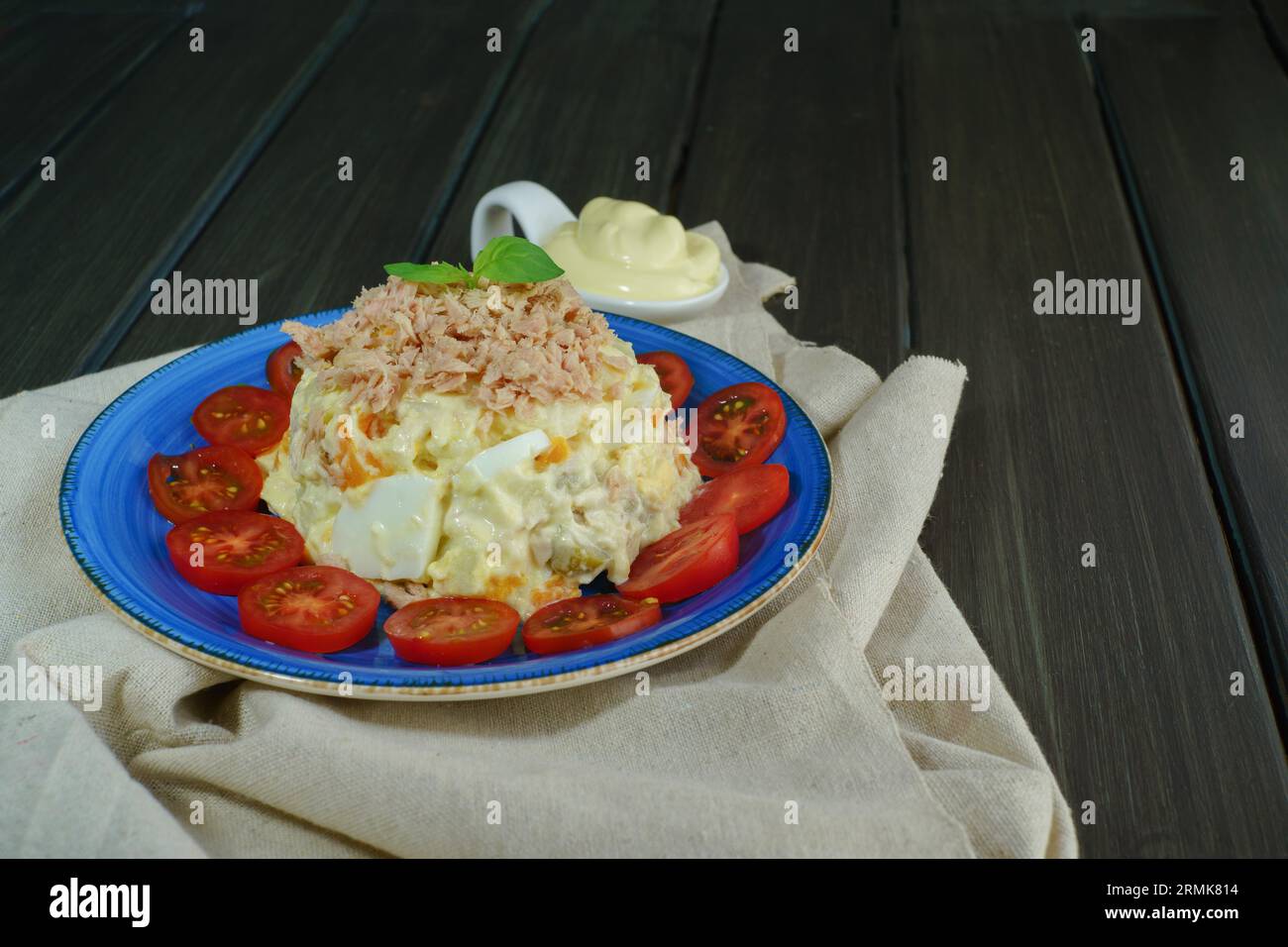 Ensaladilla rusa or russian salad, typical spanish tapa with tuna and mayonnaise on a blue plate decorated with tomato and green leaves on a raffia Stock Photo