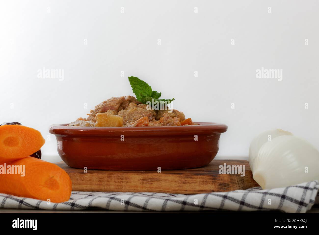 Meat stew with sauce in a clay pot on a wooden board accompanied by carrots and onions Stock Photo