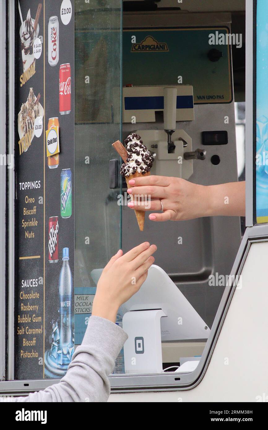 An ice cream vendor passes a 99 ice cream with added chocolate chips from the serving hatch to a waiting customer, August 2023. Stock Photo