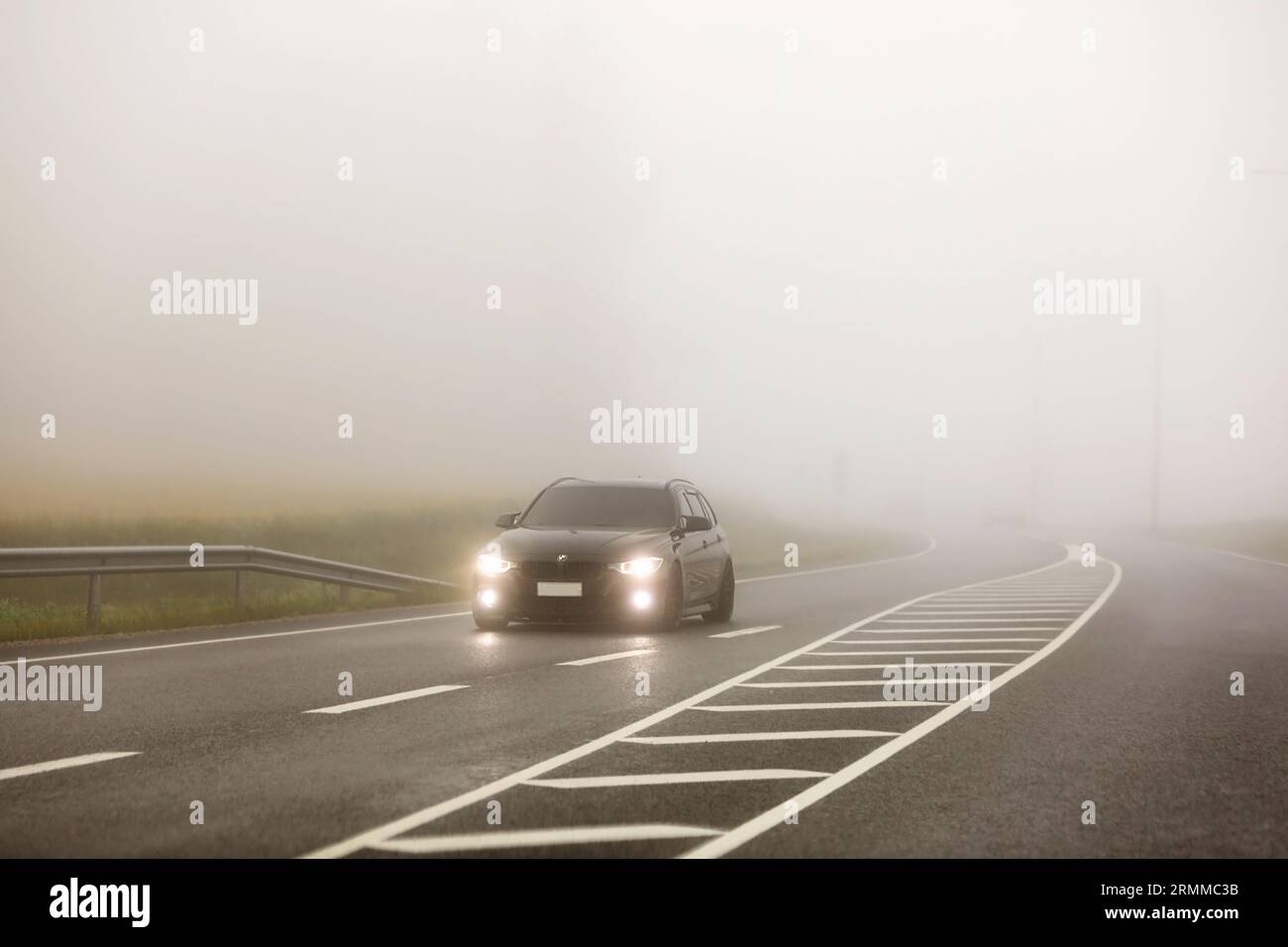 Black BMW car with bright headlights driving on road through thick morning fog. Salo, Finland. August 25, 2023. Stock Photo