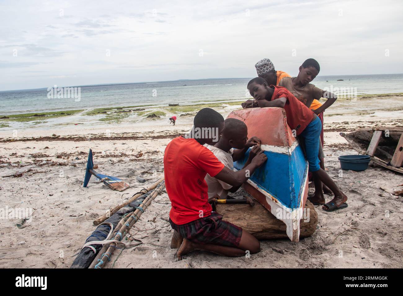Group of African people fixing and maintaining fisherman's colorful wooden boat, at the shore of Indian Ocean. Bunch of curious kids wondering around Stock Photo