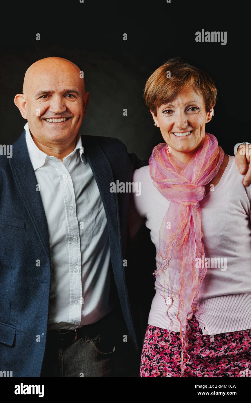 In a dimly lit studio, a long-married elder couple beams. His baldness is accentuated by a blue jacket and pale blue shirt; her attire, predominantly Stock Photo