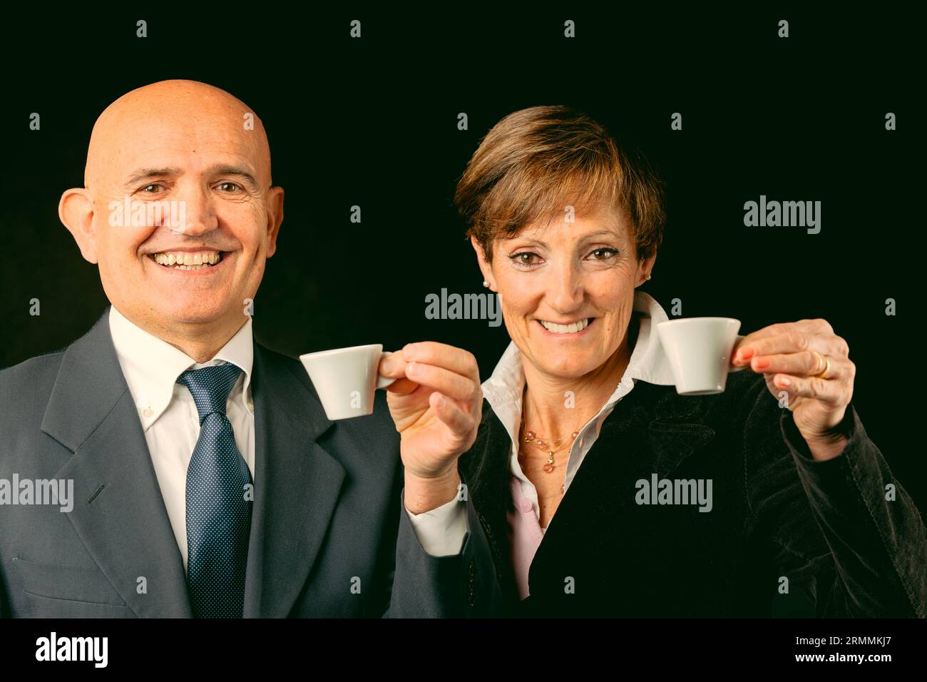 Portrayed elegantly against black, two wealthy elderly enjoy Italian espresso on their European voyage. While he's bald with jacket and shirt, she fla Stock Photo