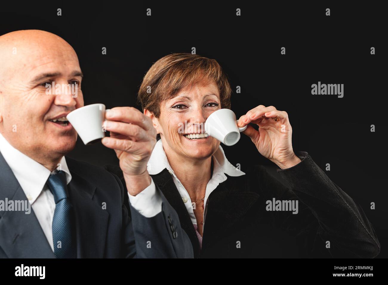 Affluent elderly duo, in peak health, savor Italian espresso amidst their European journey. Set against black, he's bald in jacket and shirt, she spor Stock Photo