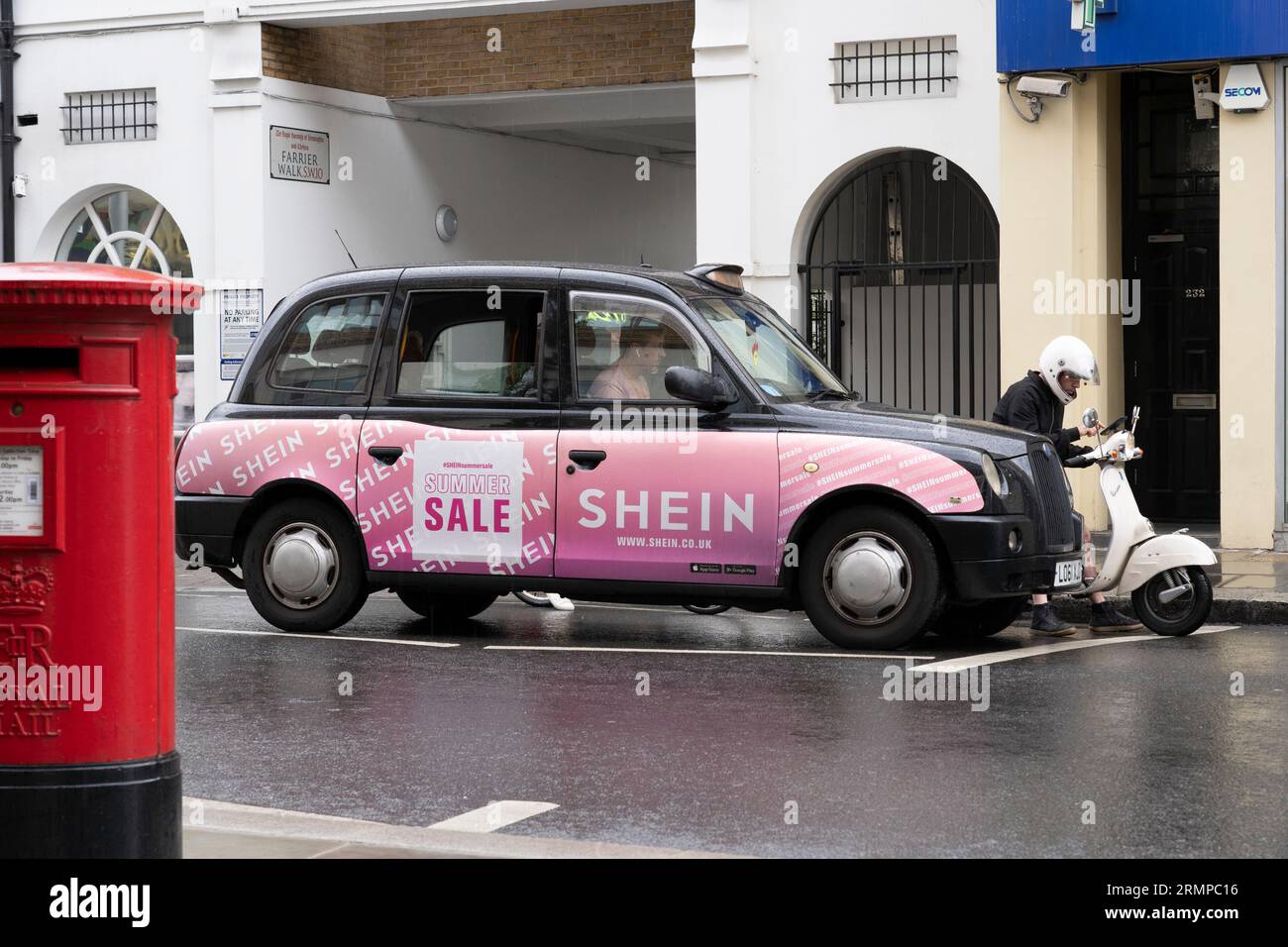 A traditional London black cab with an advertisement, next to a food delivery scooter rider, with a cyclist behind and a red pillarbox. London, UK Stock Photo