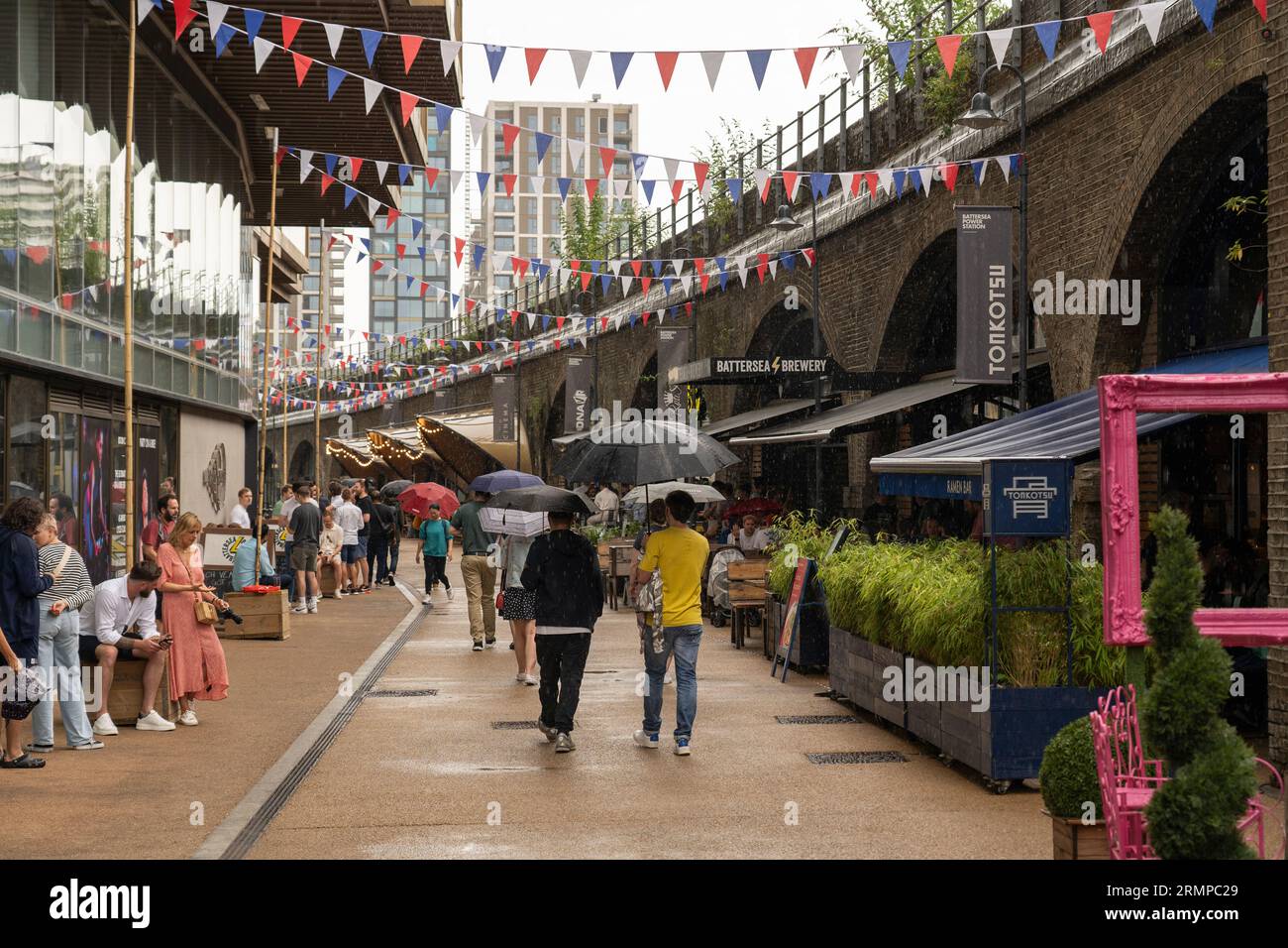 People walking in the rain and holding umbrellas on Arches Lane near Battersea Power Station next to various restaurants and bars. London, England Stock Photo