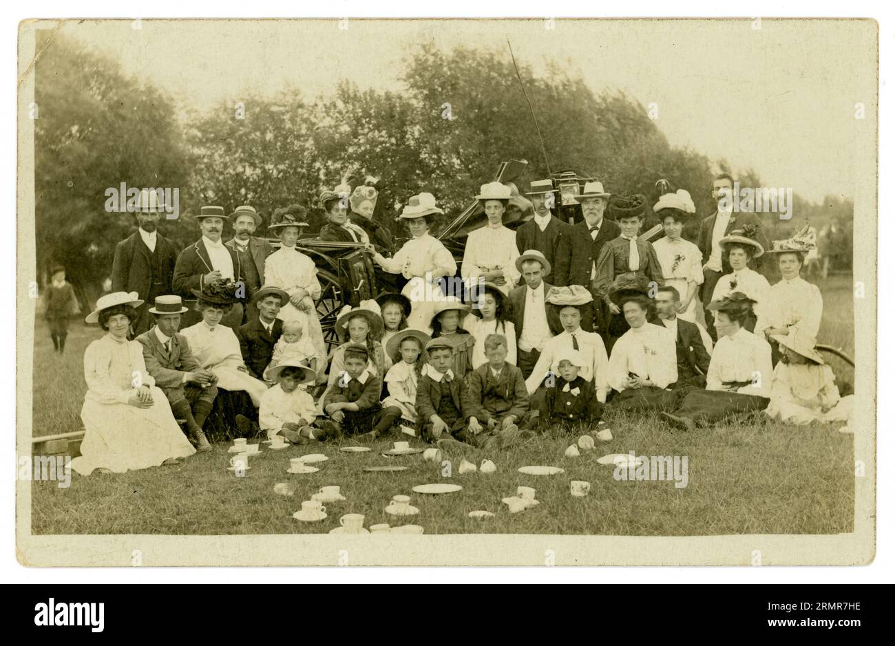 Original Edwardian era postcard of a tea party group outdoors in a field somewhere in Britain - a church outing maybe, cups and saucers strewn around, amazing selection of hats and outfits. Fascinating characters both men, women and children, even babies. A carriage is in the background with a grumpy looking older woman seated, wearing a full face veil, perhaps to keep off pesky flies. Photograph circa 1907, Possibly Newport area, Wales, U.K. Stock Photo