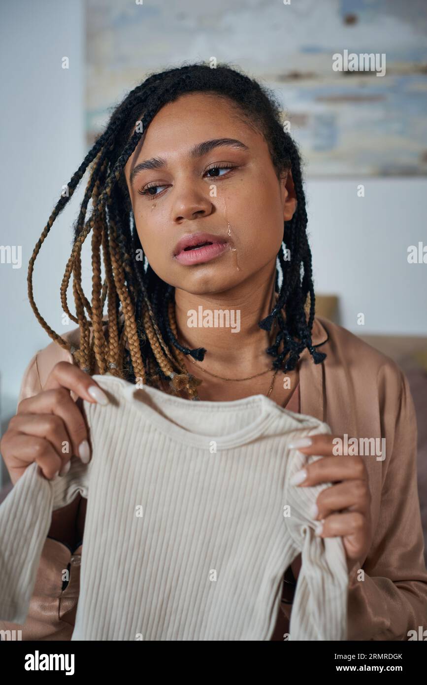 depressed african american woman holding baby clothes and crying in bedroom, miscarriage concept Stock Photo