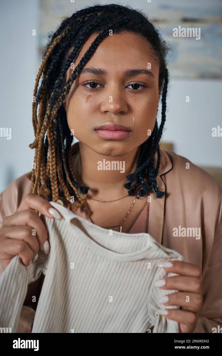 depressed african american woman holding baby clothes and crying at home, miscarriage concept Stock Photo