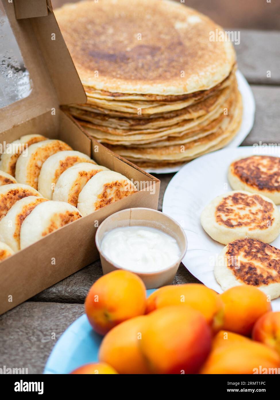 Cottage cheese pancakes, syrniki in paper box and stack thin pancakes on wood table. Delivery healthy and delicious morning breakfast. Stock Photo