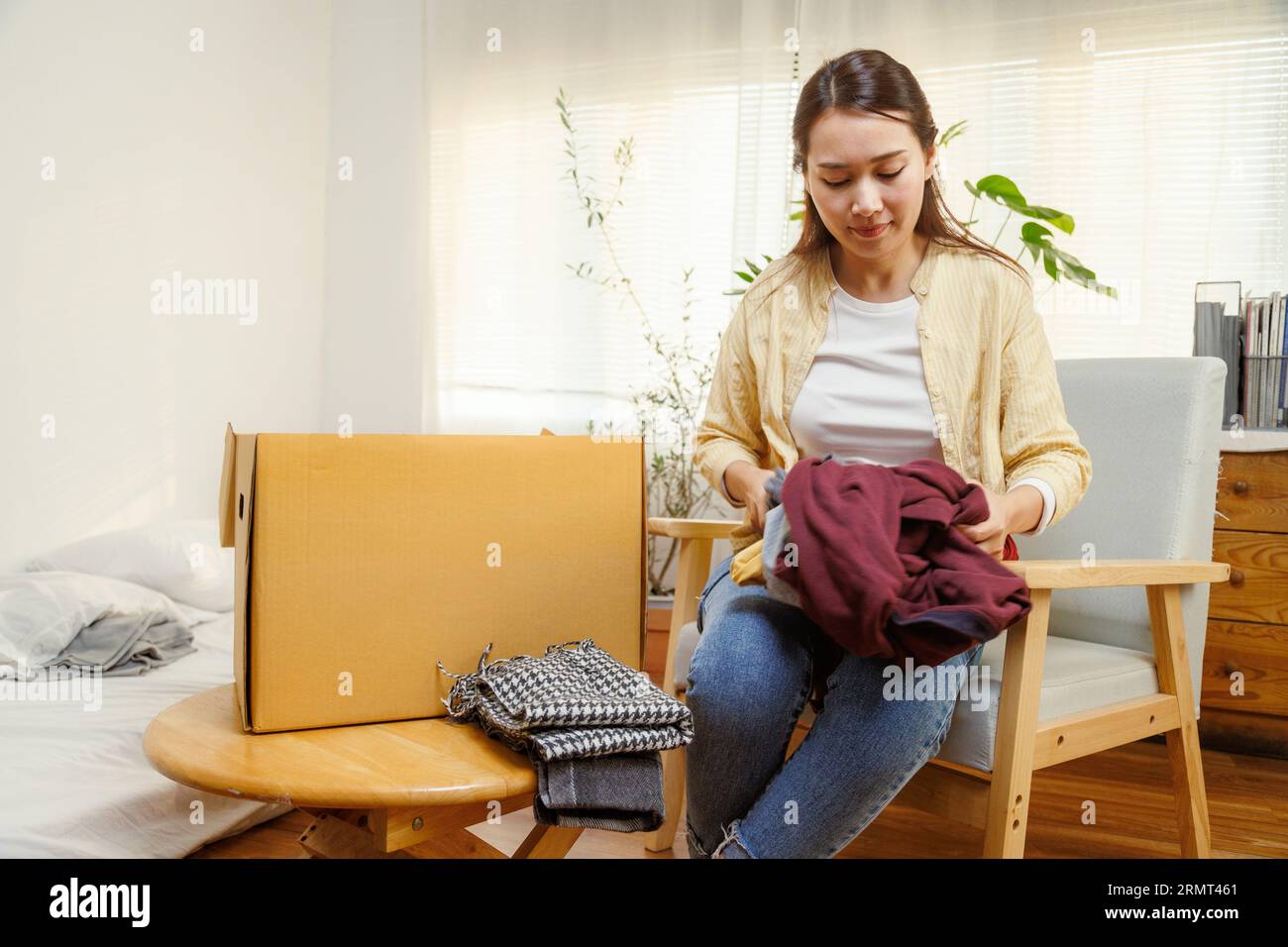 Young asian female putting clothes into cardboard box to moving to new house or apartment. Stock Photo