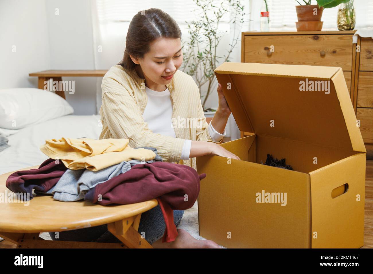 Young asian female putting clothes into cardboard box to moving to new house or apartment. Stock Photo