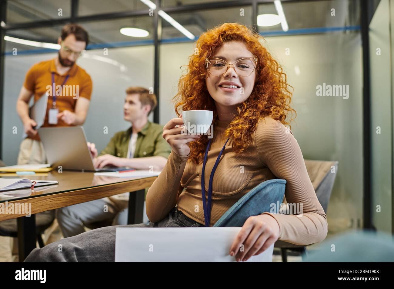 redhead businesswoman with coffee, blurred colleagues talking in office, professional headshot Stock Photo