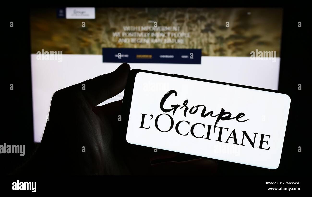 Person holding mobile phone with logo of French cosmetics company Groupe L'Occitane on screen in front of web page. Focus on phone display. Stock Photo