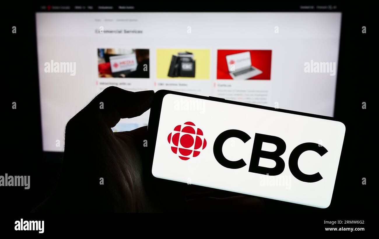 Person holding cellphone with logo of Canadian Broadcasting Corporation (CBC) on screen in front of webpage. Focus on phone display. Stock Photo