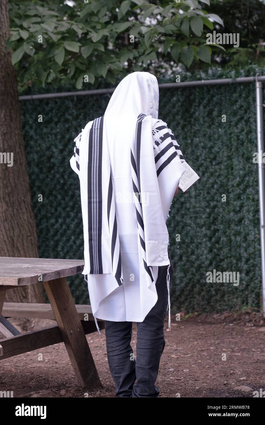 An orthodox Jewish man photographed from behind, reciting morning prayers outdoors between trees