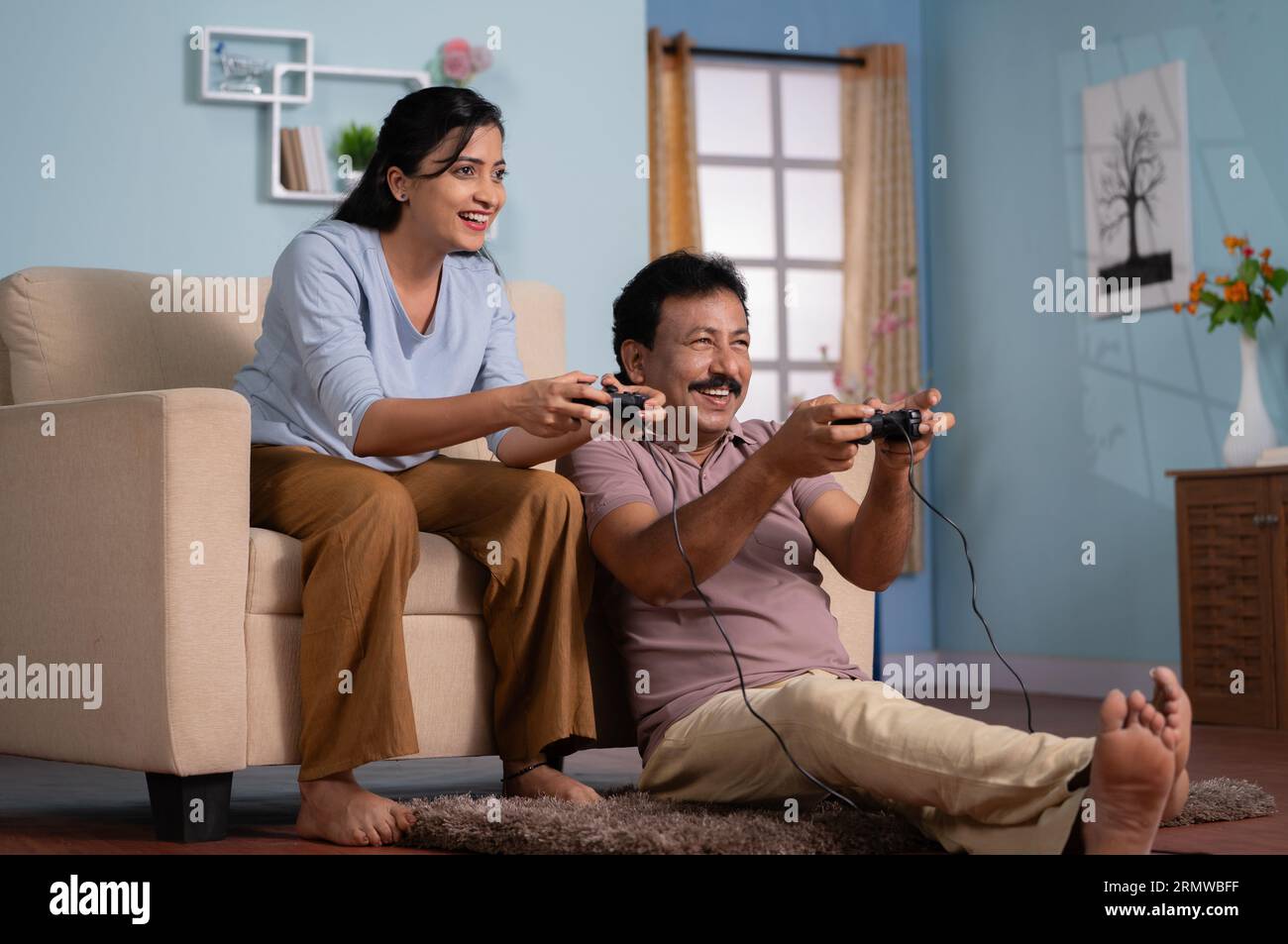Cheerful indian father with adult daughter playing video game by using joystick at home - concept of entertainment, leisure activity and family Stock Photo