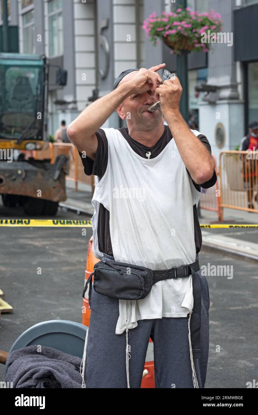 A Jewish man wearing a skullcap and tzitzits blows a shofar on 8th Avenue & 34th Street in Manhattan, New York City. Stock Photo