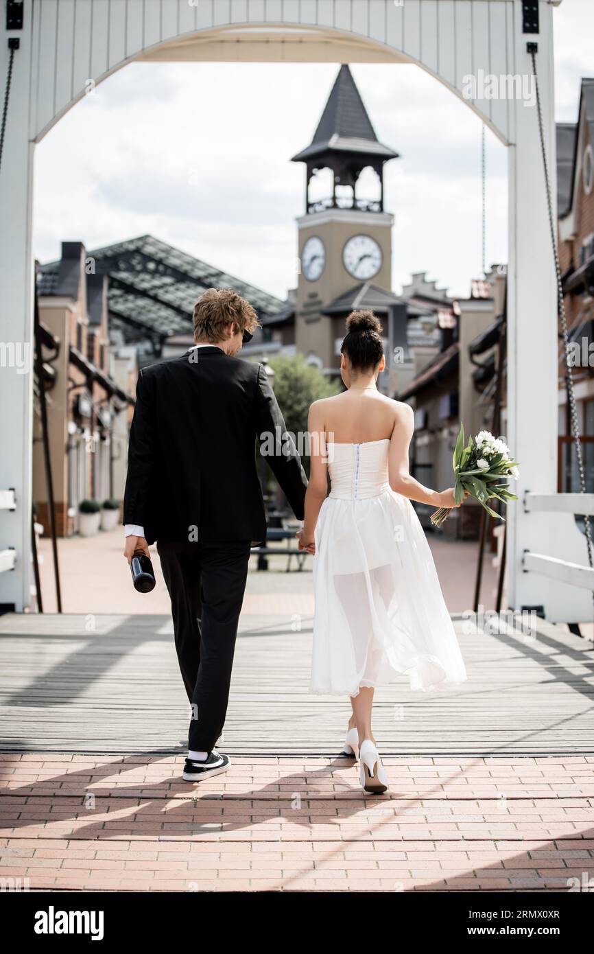 back view of romantic multiethnic couple holding hands and walking on ...