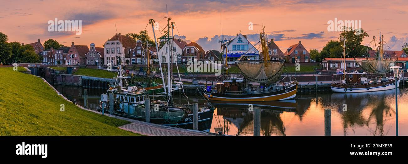 A wide 3:1 panorama photo of the picturesque harbor in the fishing village of Greetsiel after sunset. Greetsiel is a small harbor town on the Leybocht Stock Photo