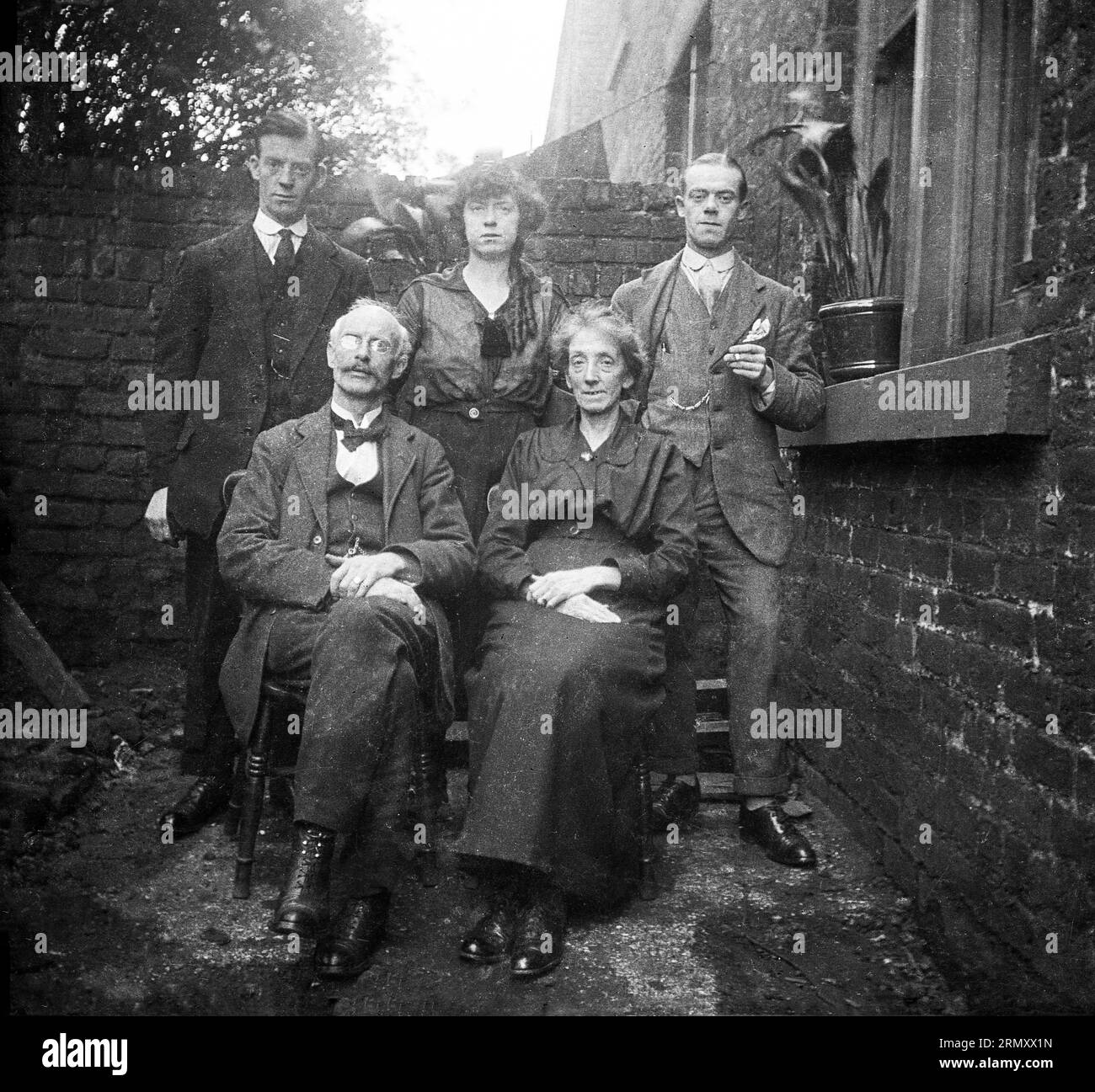 1930s, historical, a family photo outside in an alley of a victorian house, England, UK. The men and ladies are wearing the formal clothes of the era, including gold chains, stiff collar and laced up over ankle boots worn by the elderly gentleman sitting at the front. Stock Photo