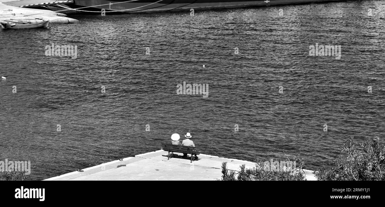 A grayscale shot of an old couple sitting on a bench near a lake Stock Photo